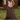 A woman stands outdoors in front of a rustic red wooden shed, wearing a cream ribbed tank top, a long brown and black plaid skirt, and a pink knit beanie.