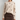 A woman wearing a cream-colored fuzzy knit sweater with small dark brown heart appliqués, paired with a brown button-front skirt, against a plain light gray studio background.