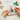 A person's hands are shown cross-stitching a colorful cactus design in a wooden hoop. The scene includes an embroidery kit card, a finished sample print, and threads on a white desk, making it a Lifestyle shot.