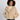 A woman wearing a beige curly sherpa fleece jacket over a white lace top and dark brown jeans, standing against a light gray studio background.