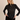 A blonde woman wearing a black sheer lace long-sleeve top with a textured floral pattern and black satin pants, posing against a solid light beige background.