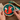 A person holds a circular embroidery hoop with a partially finished fox needlepoint or cross-stitch design in a wooden frame. The design shows an orange fox with white and cream accents on a dark teal background with green leaves and small yellow stars.